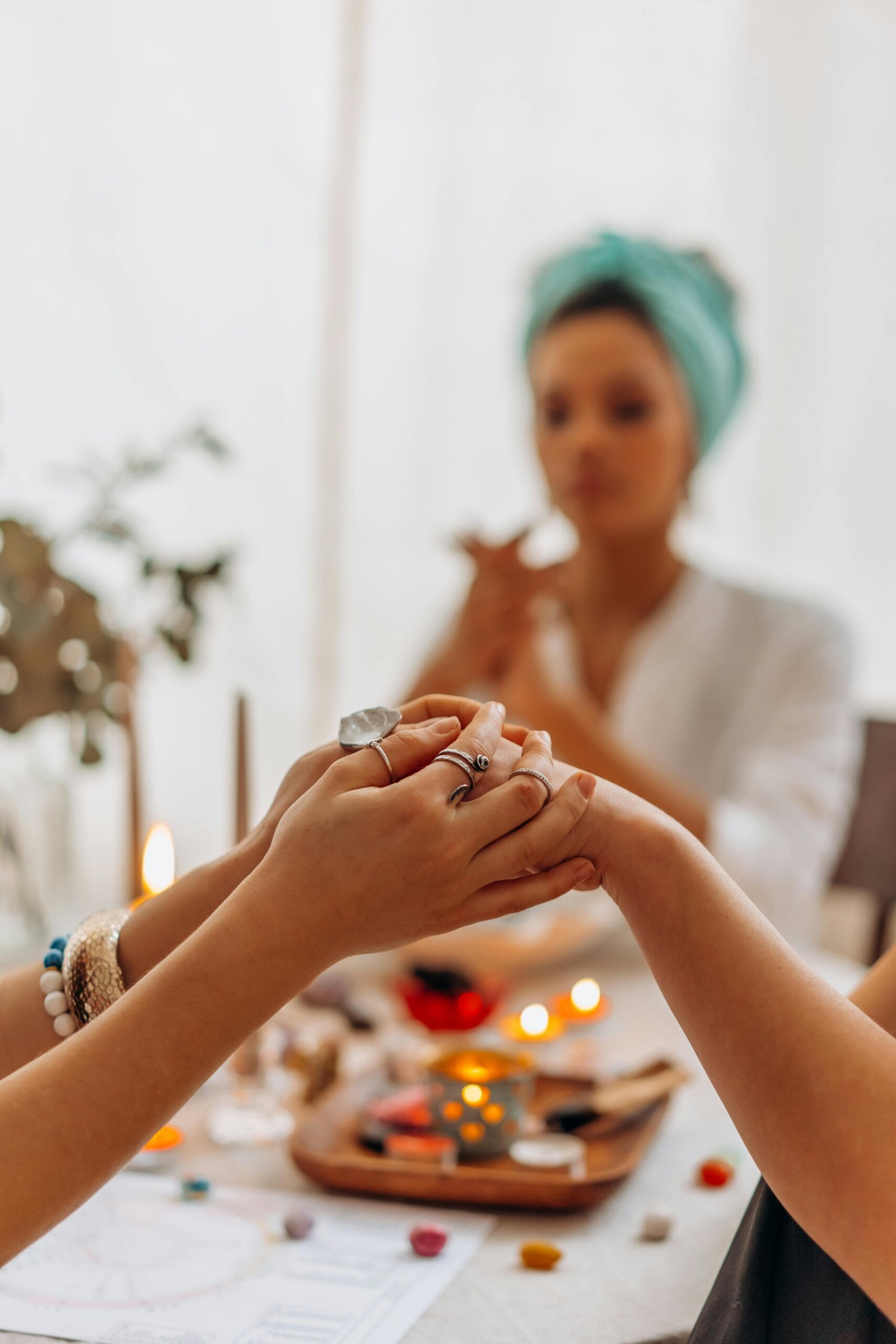 The image depicts a serene and spiritual setting where two individuals are engaged in a healing practice, possibly involving energy work or meditation. In the foreground, we see one person holding the hands of another, with a focus on their hands adorned with rings and bracelets. In the background, there is another person who is holding a crystal or incense, surrounded by soft candlelight, creating a peaceful atmosphere. The table is adorned with crystals, stones, and a chart, likely related to a holistic or spiritual practice.