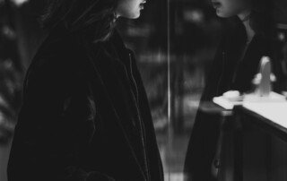 A black and white image of a woman looking at herself in the mirror and her reflection showing in the other side of the mirror. This represents the topic of blog that talks about the shadow self