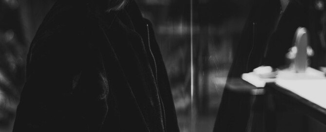 A black and white image of a woman looking at herself in the mirror and her reflection showing in the other side of the mirror. This represents the topic of blog that talks about the shadow self