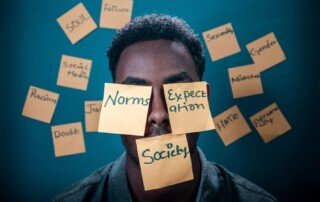 An image of a man whose face especially lips and eyes are covered with post it notes that says society, expectations, norms, showing how sometimes we are unable to speak our truth and close our eyes due to external factors