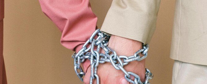 A man and a woman both holding hands tied together with a metal chain showing they are stuck in a toxic pattern