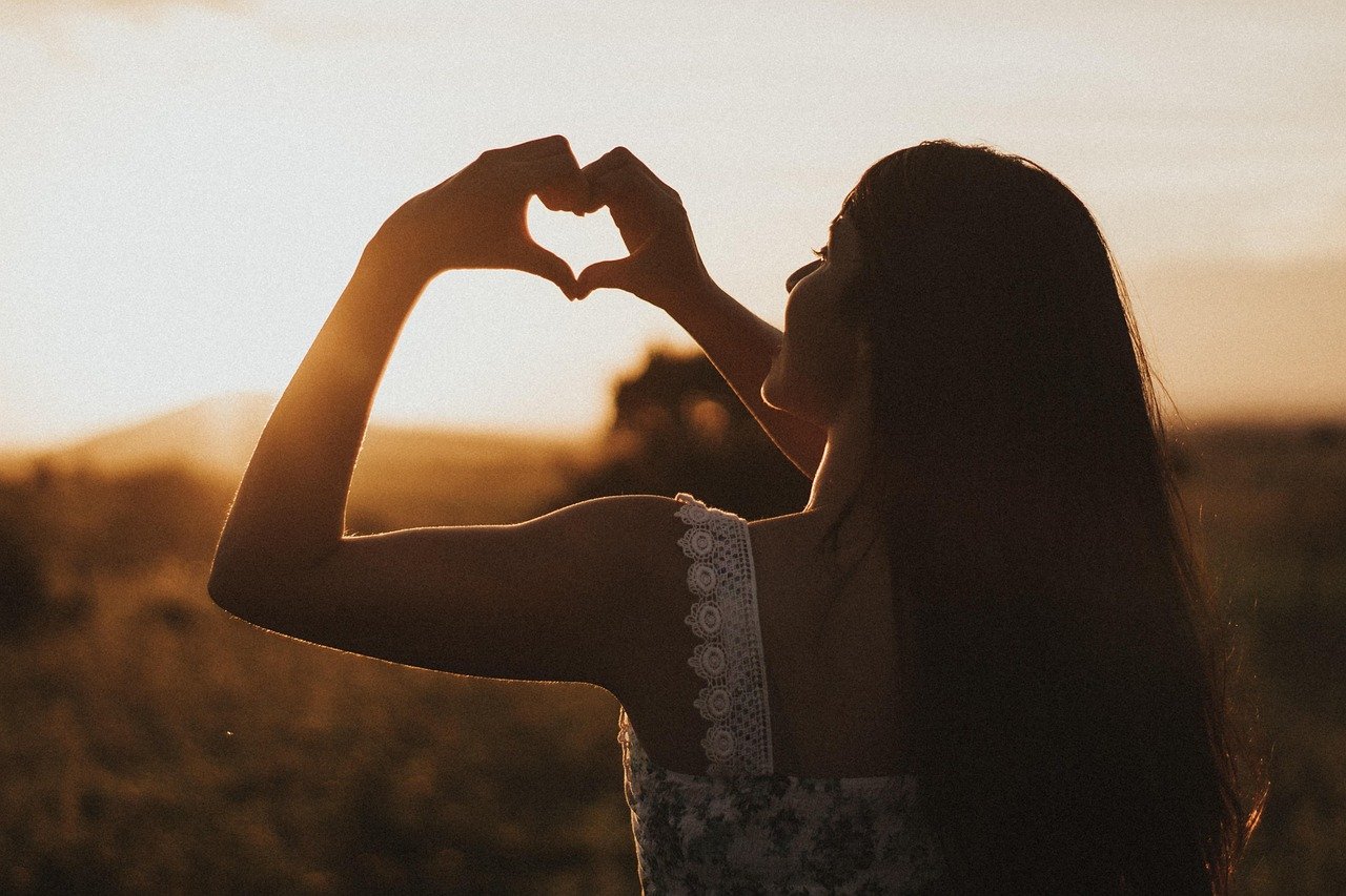 Is thetahealing a religious practice **Alt text:** A woman standing outdoors at sunset forms a heart shape with her hands in front of the sun. She has long dark hair and is wearing a sleeveless floral dress with lace straps. The warm light creates a silhouette effect, highlighting a peaceful and loving moment in nature.