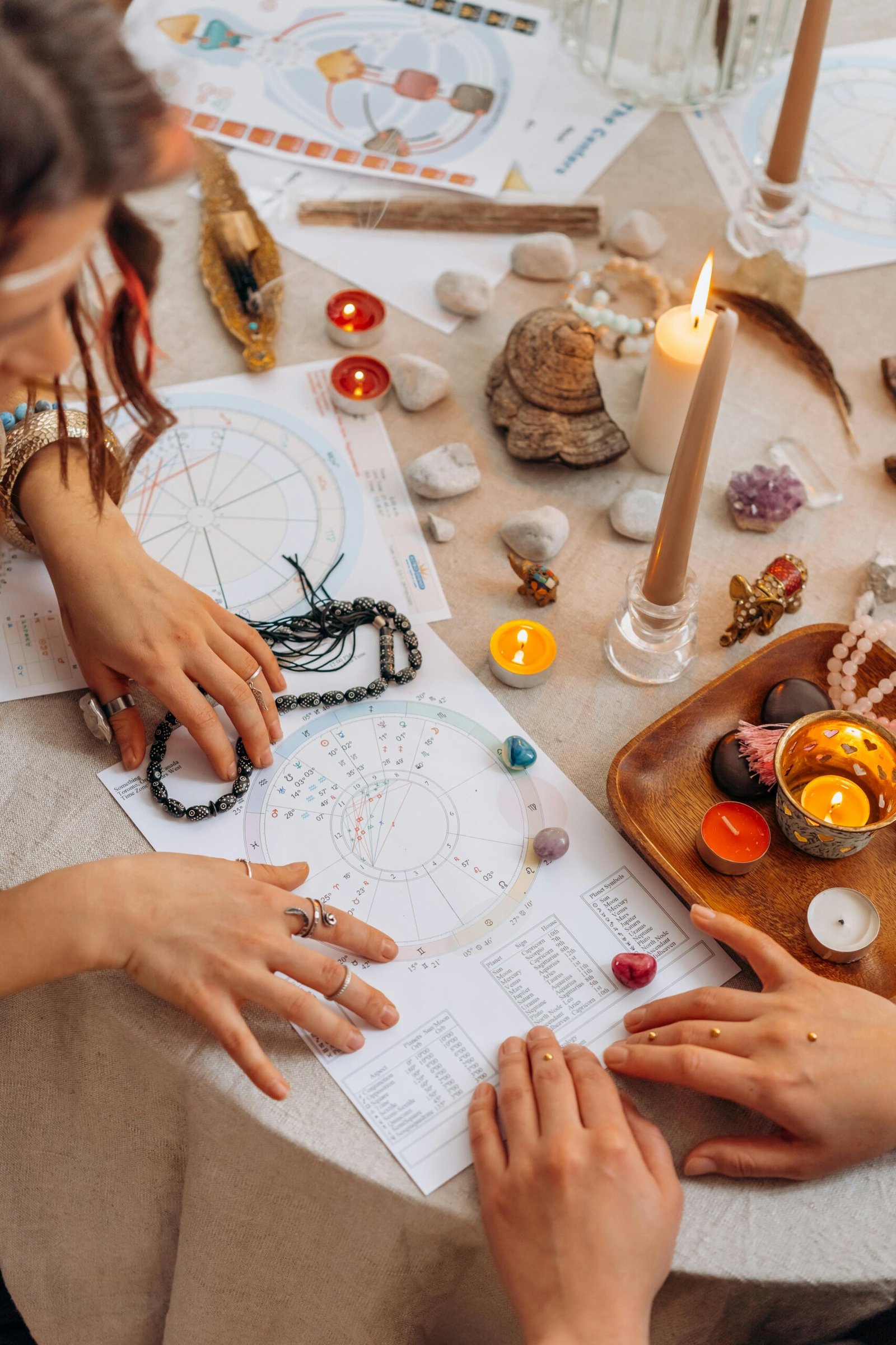 Spirituality and thetahealing A close-up of a spiritual or astrology-themed setting with two people examining astrological birth charts on a table. The table is decorated with candles, crystals, stones, incense, beads, and symbolic items, creating a mystical and ritualistic atmosphere. One person is holding a beaded necklace, and both have adorned hands, suggesting an intentional or ceremonial practice.