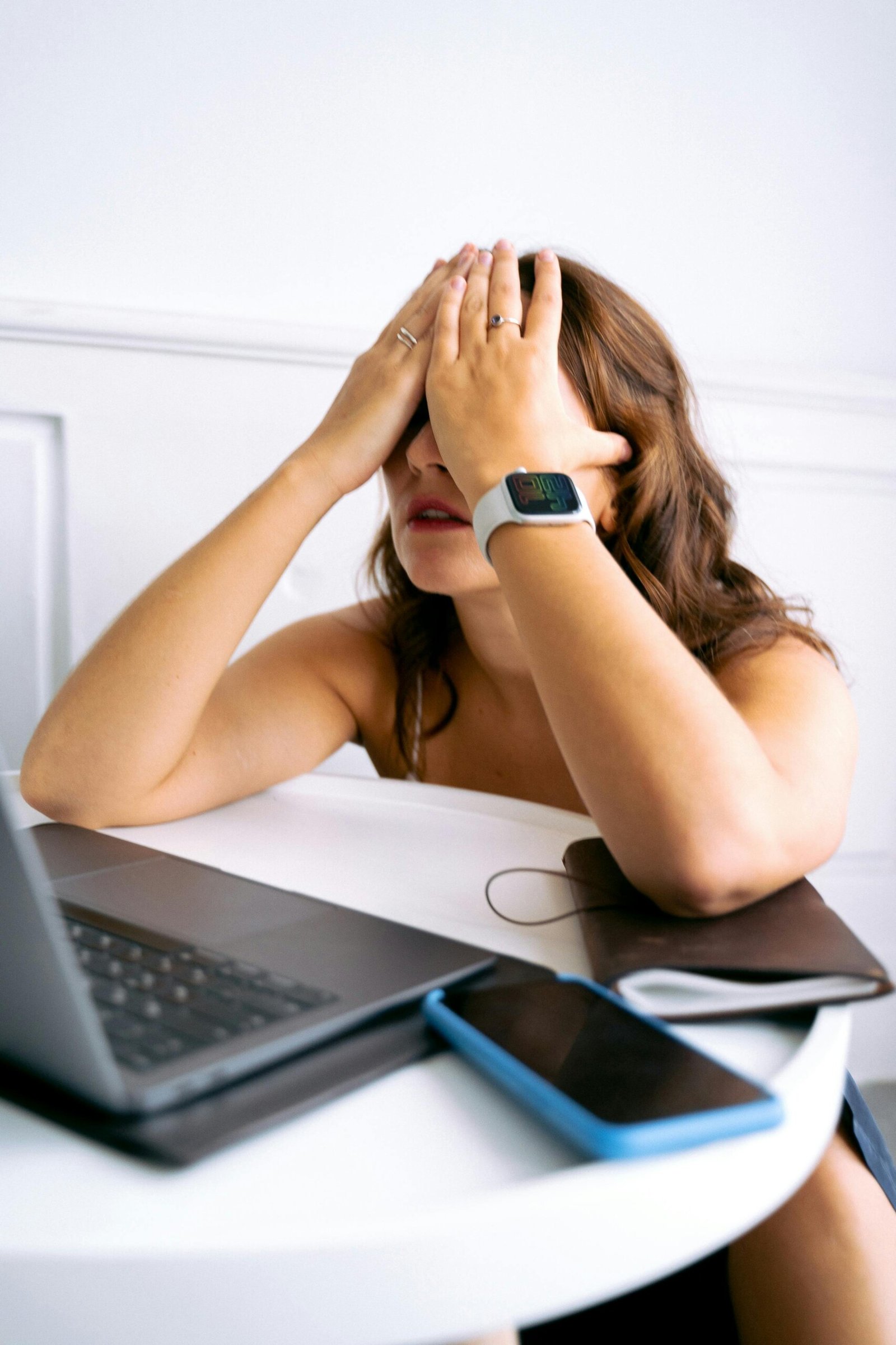 Who can practice thetahealing A woman sitting at a white table appears stressed or overwhelmed, covering her face with both hands. She is wearing a smartwatch and has a laptop, smartphone, and notebook in front of her, suggesting a work or study-related situation. The background is minimal and white, drawing focus to her emotional state.