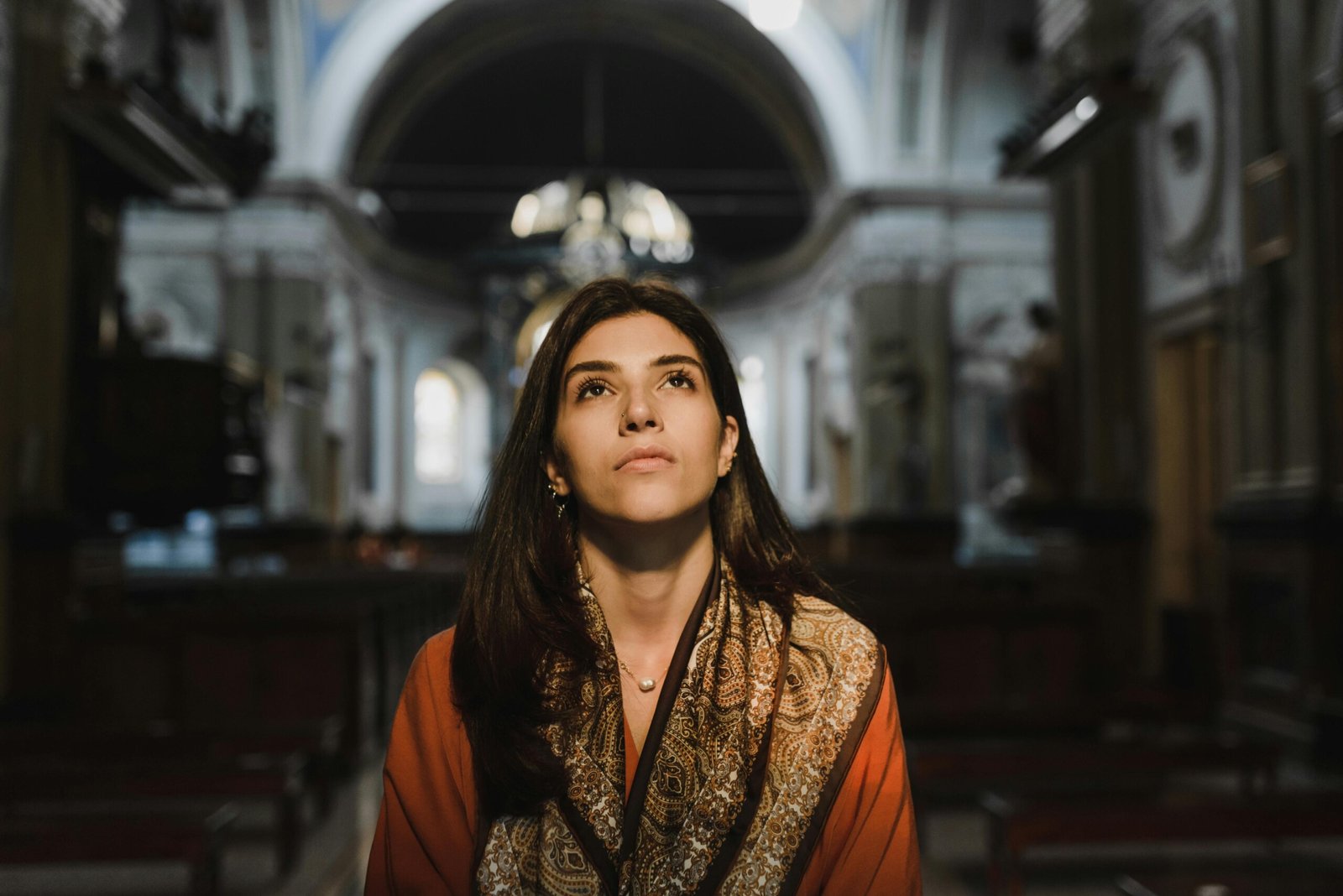 feeling non intuitive A woman with long dark hair and a patterned scarf sits in a dimly lit religious place, looking upward with a thoughtful or contemplative expression. The architecture and soft light from the arched windows create a serene and reflective atmosphere.